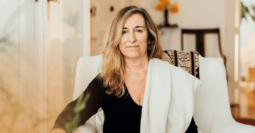 Woman sits in a white chair staring straight at the camera. Her facial expression is interested. She is wearing a black long sleeve shirt. The background is a dark brown wooden chair against a white wall.