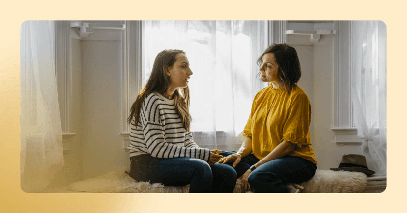 A mother and daughter sit on a bench in front of a window in serious conversation. The daughter is wearing a black and white striped shirt and the mother is wearing a yellow sweater The image is framed in yellow.