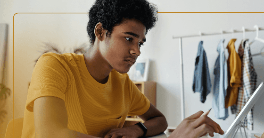 Young man sits at a desk staring at a tablet in front of him. His facial expression is serious as he scrolls and holds a pencil with his right hand. He is wearing a yellow long sleeve shirt. The background is a white wall with clothes hanging on a rack in front of it.