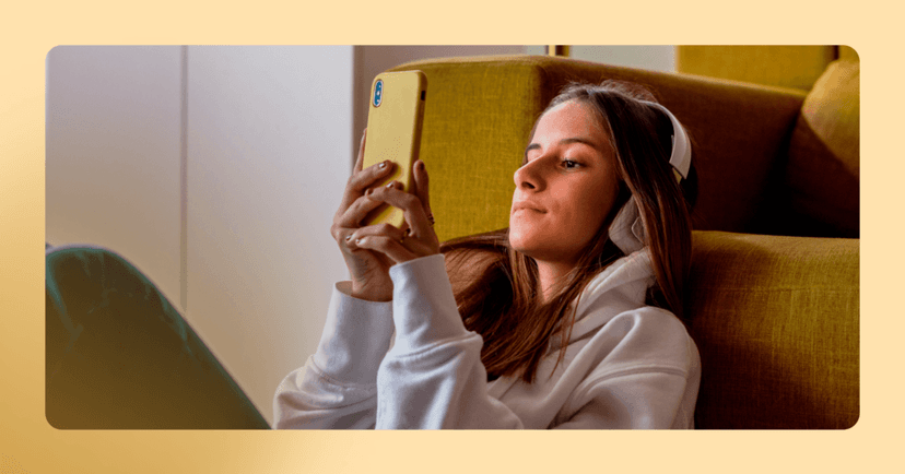 Young girl sits on the floor leaning against a yellow couch. She is looking at the yellow phone she holds in front of her in both hands. She is wearing white over the ear headphones and a white hoodie. There is a yellow border around the image.