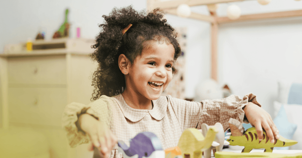 A young girl in a play room with a big smile playing with dinosaur toys.