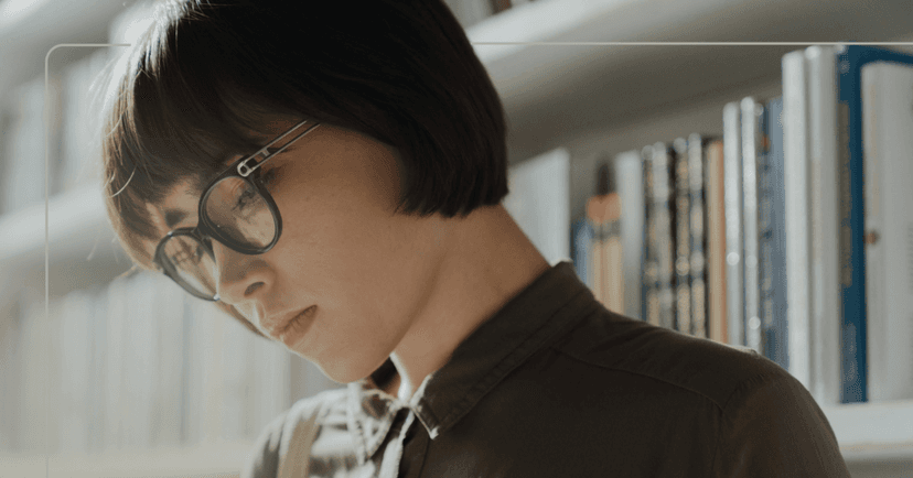 Woman stands in front of a book shelf. She is looking down at something in front of her offscreen. She is wearing black round glasses and a brown button-up shirt. The background is hardcover books that are blurry.