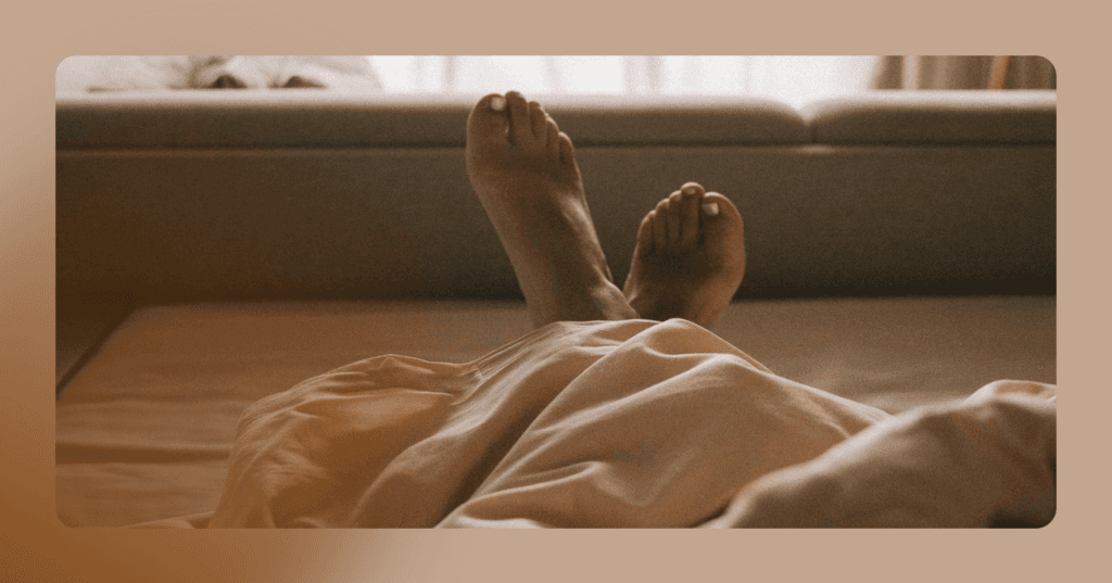 Image shows feet crossed over each other coming out of a sheet. The upper-half of the individual is not visible. The person is lying down in bed. The background is a couch.