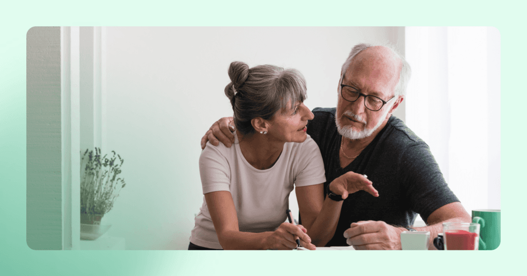 Couple is talking at the kitchen counter table. Woman is looking at the man on her left with a pen in her left hand. Man is sitting with his right arm around her shoulder. She is wearing a light pink T-shirt and he is wearing a gray V-neck. The background is a white wall.