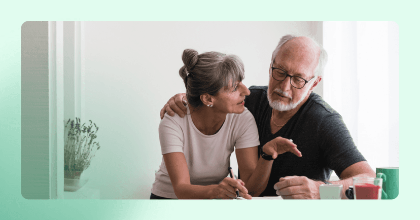 Couple is talking at the kitchen counter table. Woman is looking at the man on her left with a pen in her left hand. Man is sitting with his right arm around her shoulder. She is wearing a light pink T-shirt and he is wearing a gray V-neck. The background is a white wall.