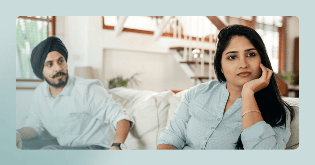 Couple sits on a couch downstairs in their home. The man is on the left looking at the woman. His facial expression is angry. The woman is on the right leaning on her left hand staring off. She is wearing a blue button-up blouse and is sharper in focus.