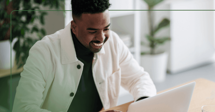 Man in a white button down shirt looks down at a laptop while smiling. He is sitting at a wooden table with plants of our focus in the background.