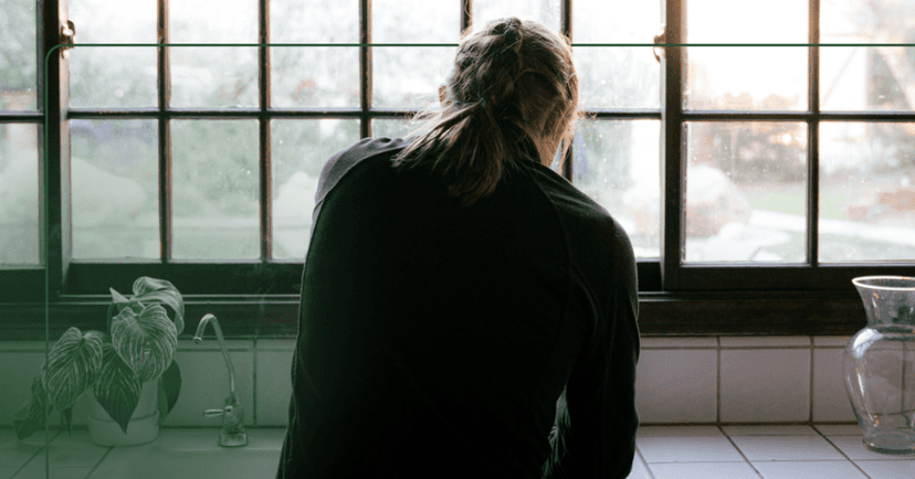 A woman in a dimly lit kitchen washes dishes at the kitchen sink. Her back is facing the camera and she is wearing a black half zip. There is a plant on the kitchen counter and the kitchen windows overlook a front yard.