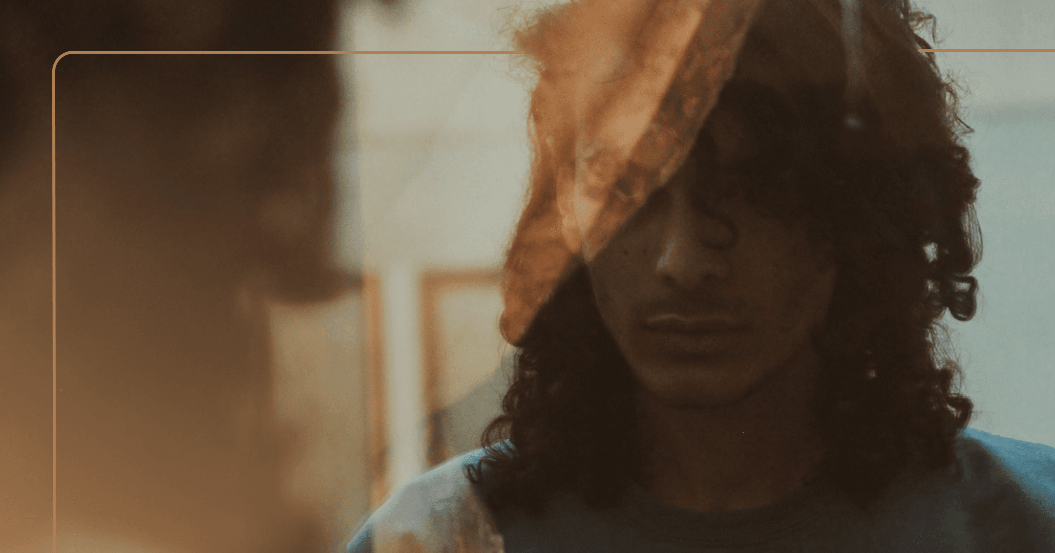 A man with long, dark curly hair looks at himself with a serious, somber look on his face. His hair is partially blocking his eye. The image is framed with a yellow outline.