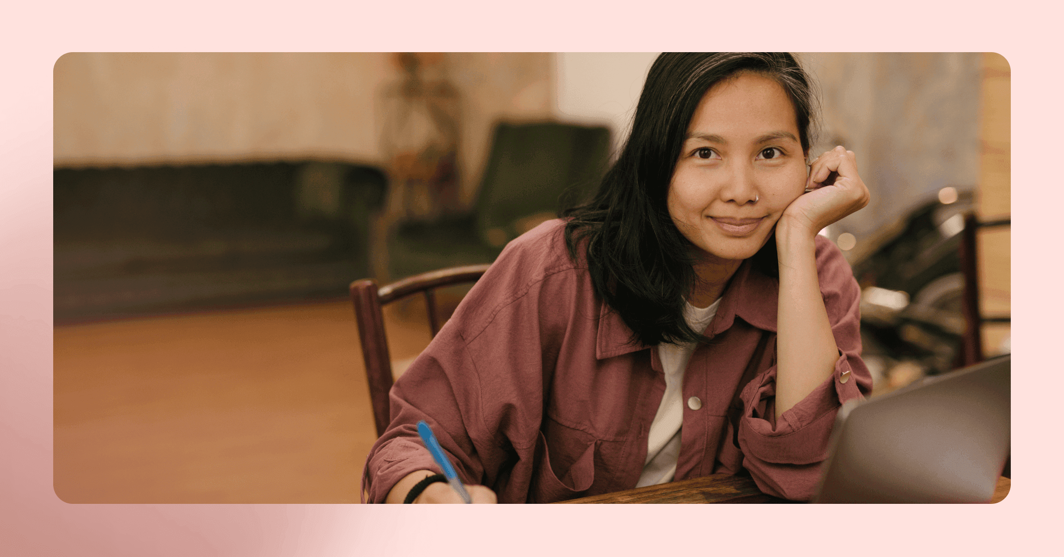 Woman in a maroon button up shirt sits at a wood table and stares at the camera while leaning on her hand. She is sitting on a wood chair with a couch out of focus in the background. The image is framed in pink.