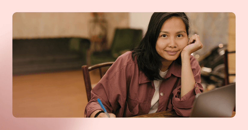 Woman in a maroon button up shirt sits at a wood table and stares at the camera while leaning on her hand. She is sitting on a wood chair with a couch out of focus in the background. The image is framed in pink.