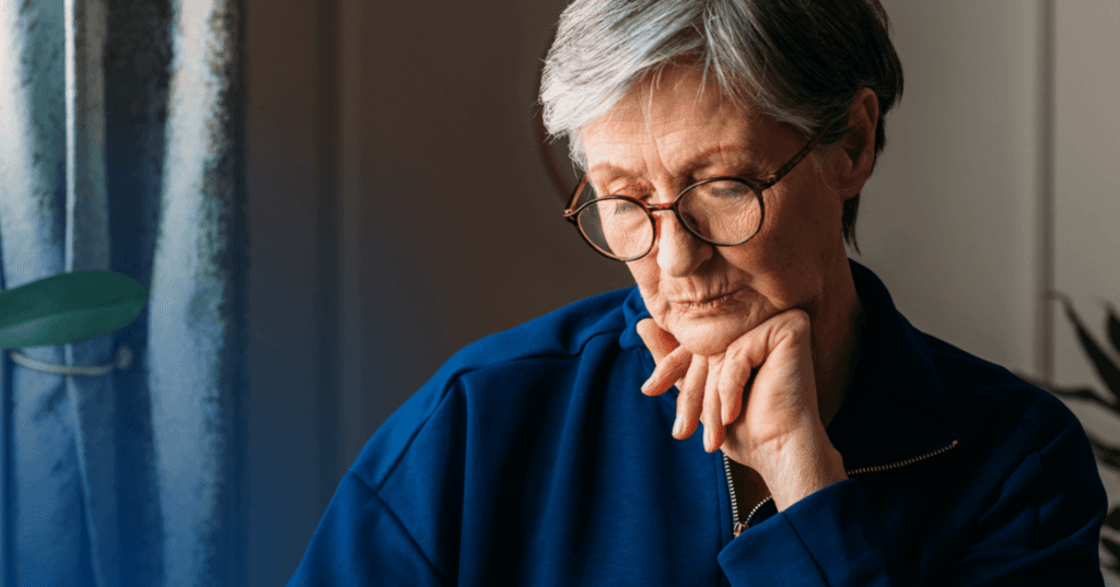 Woman sits with her left hand underneath her chin. Her eyes are closed and lips are pursed. She is wearing a blue jacket and glasses. The background is a white wall and pulled back curtains on the left side of the screen.