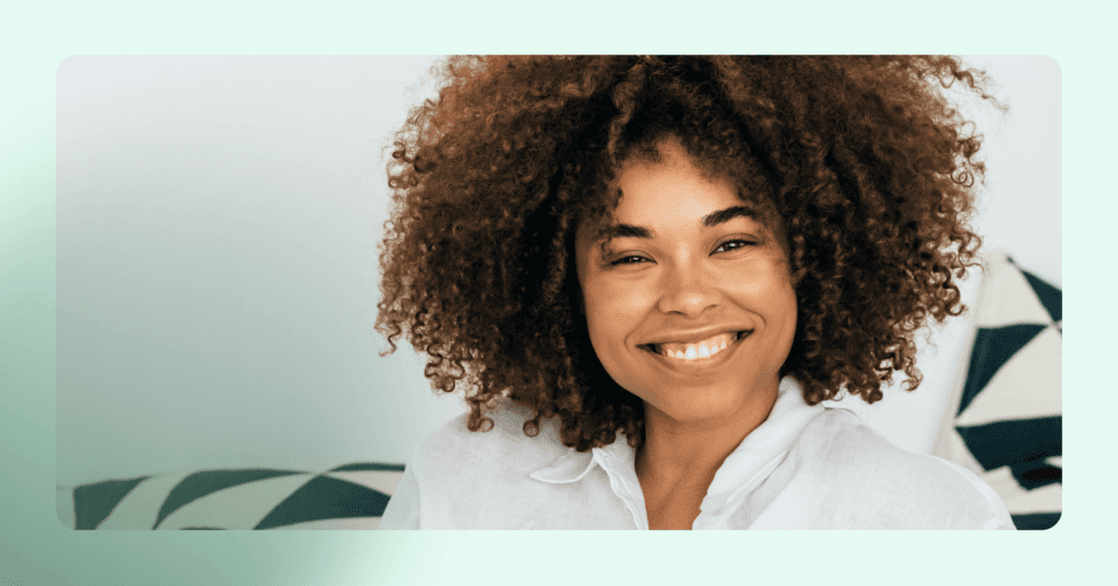 Woman is staring straight into the camera smiling with her teeth showing. She is wearing a white button up shirt. The background is a white wall with two patterned pillows.