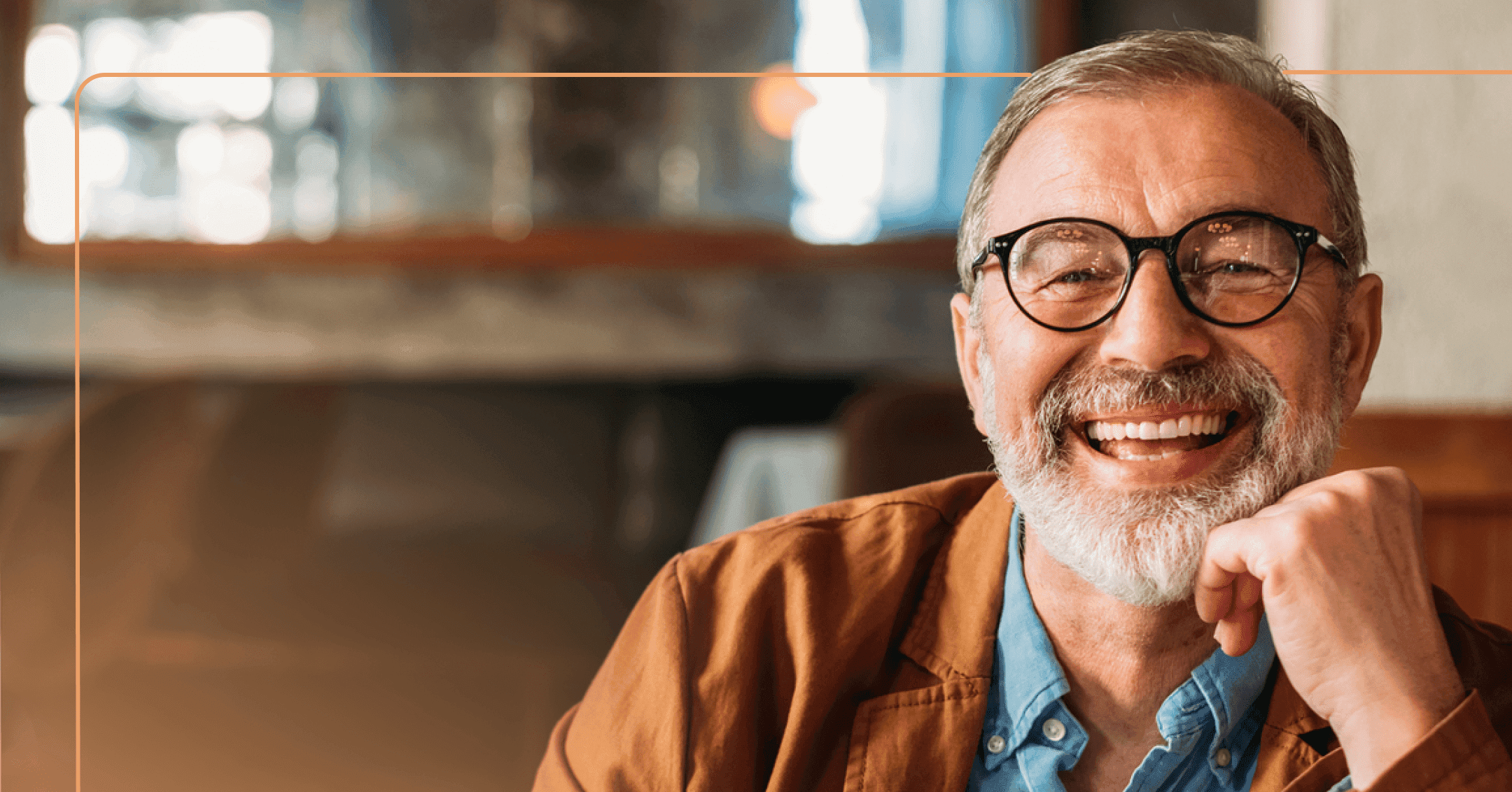 A man with a beard and glass looks directly at the camera with a big smile. He is wearing an orange jacket and a denim shirt.
