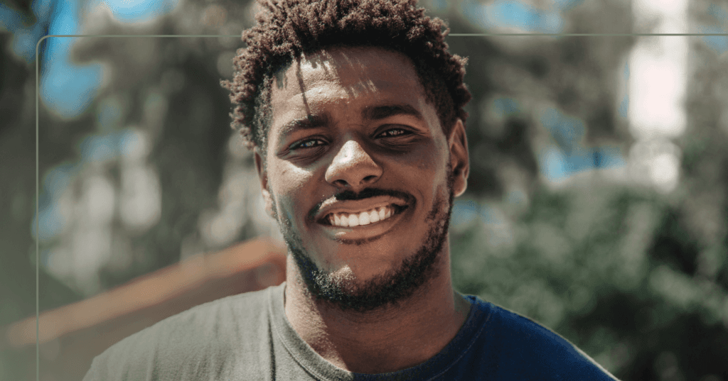 Man stands outside staring straight at the camera. He is smiling with all his teeth and the sun is shining. He is wearing a black T-shirt. The background is trees that are blurry.