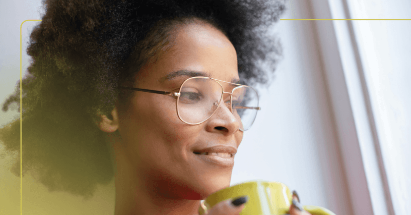 Woman stands staring out the window to her left. Her facial expression is calm and her teeth are visible in an almost smile. She is wearing glasses and holding a yellow green mug. The background is a white wall.