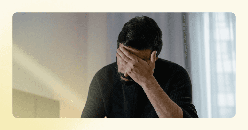 Man sits on the edge of a bed with his face in his left hand. His facial expression is not visible. He is wearing a black long sleeve shirt. The background is light blue curtains on a window.