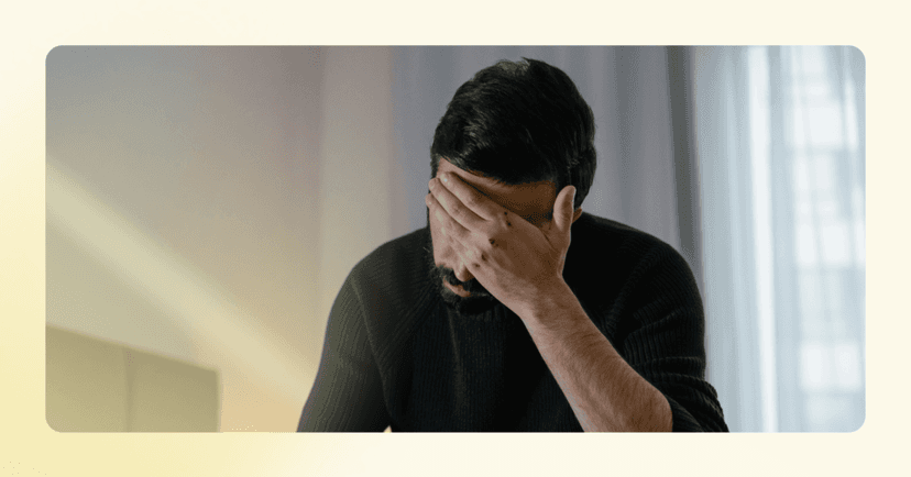 Man sits on the edge of a bed with his face in his left hand. His facial expression is not visible. He is wearing a black long sleeve shirt. The background is light blue curtains on a window.
