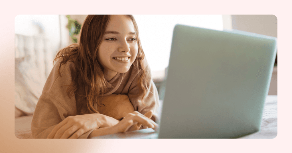 Young girl is lying on her stomach on her bed scrolling on her laptop. She is smiling and staring at the laptop screen. She is wearing a brown pullover sweater. The background is a white bedroom.