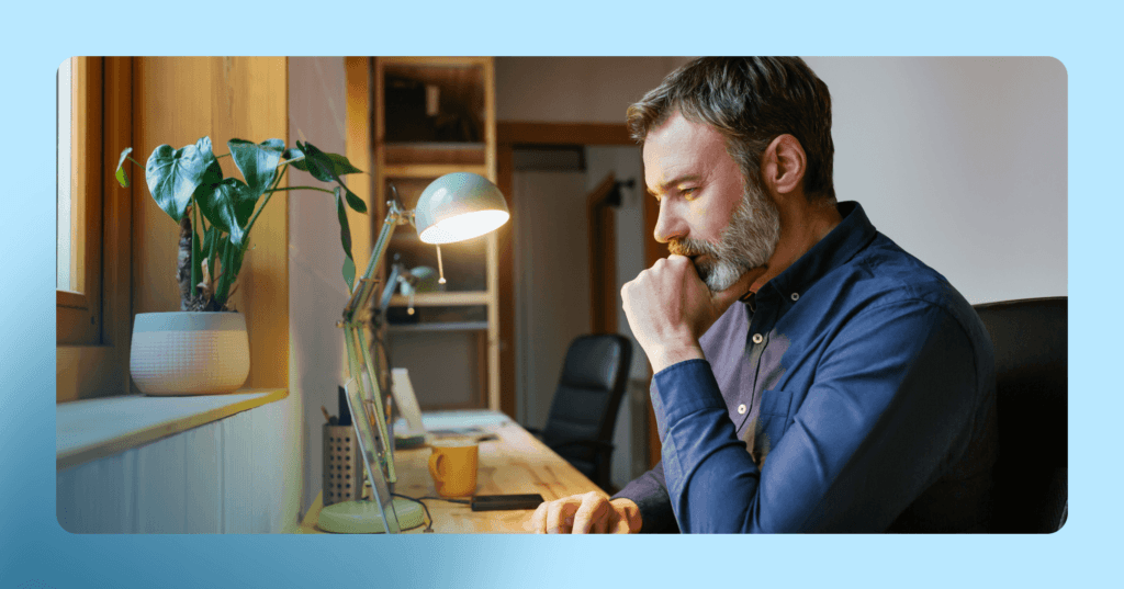 Man in a blue shirt sits at a wooden desk while working on a laptop. There is a desk light on and a plant on a shelf in the background. The image is framed in blue.