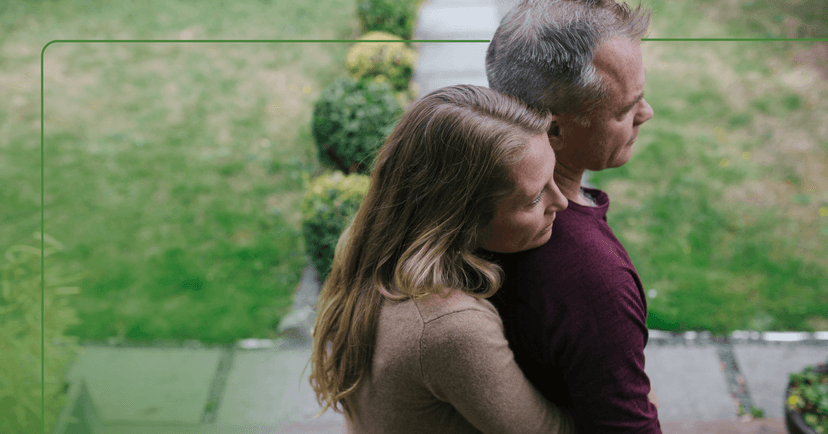 A man is embraced by a woman as they both look off camera to the right. They are standing outside on a sidewalk and grass is visible in the background.