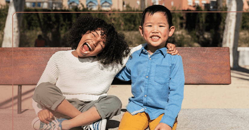 Two children sit on a park bench with their arms around each other. They are both smiling with their teeth. The girl is wearing a white sweater, green pants, and blue shoes. The boy is wearing a blue button-up shirt and yellow pants. The background is trees.