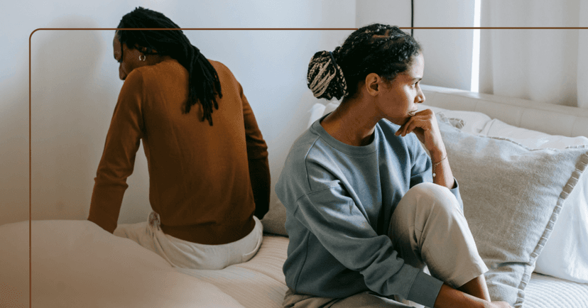 Couple sits together on opposite sides of a bed. The woman is leaning on her left hand staring out the window to the left. The man is facing the wall but looking down at the floor. She is wearing a blue sweatshirt. He is wearing a brown long sleeve shirt. The background is white curtains.
